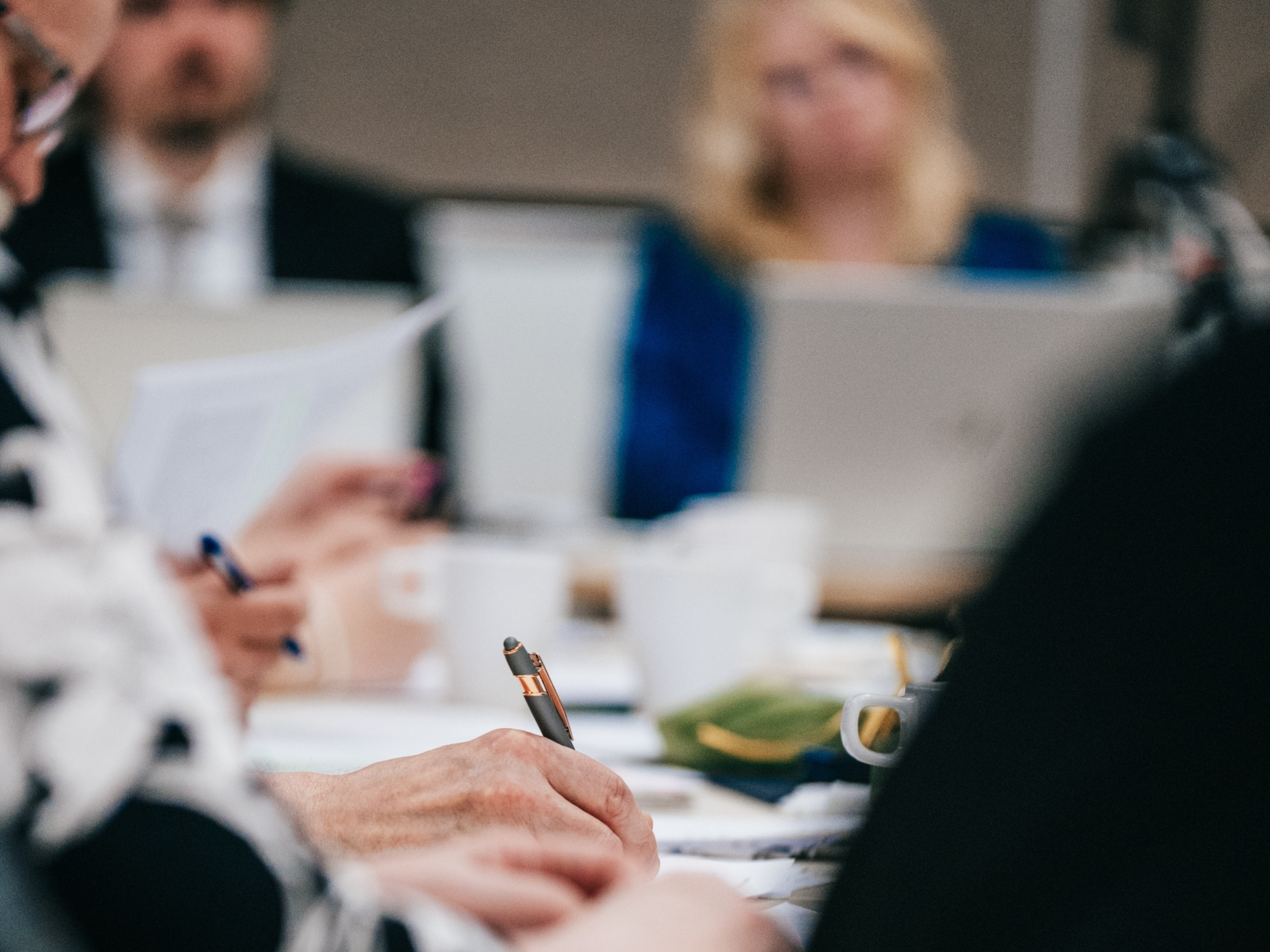 Kokouksen osallistujia näkyy sumeasti kuvan taustalla, etualalla käsi tekee muistiinpanoja.