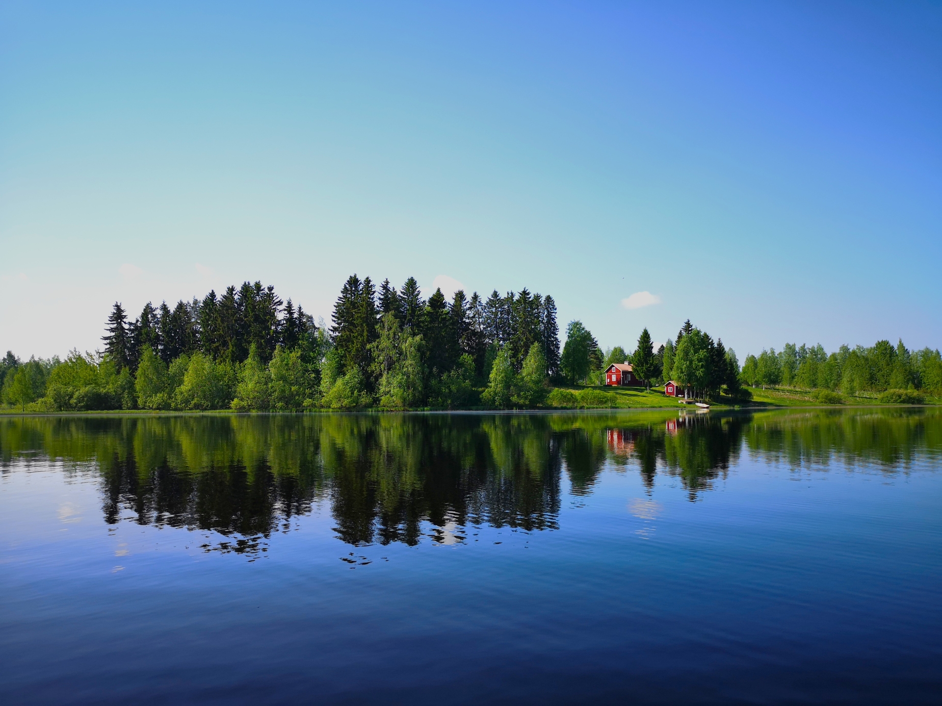 Punainen tupa saaressa ja tyyntä järvenpintaa aurinkoisena kesäpäivänä.