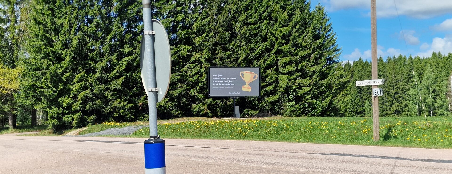 Digitaalinen infonäyttö ja siinä esitettävä mainos kuusiaidan edessä aurinkoisena kevätpäivänä.