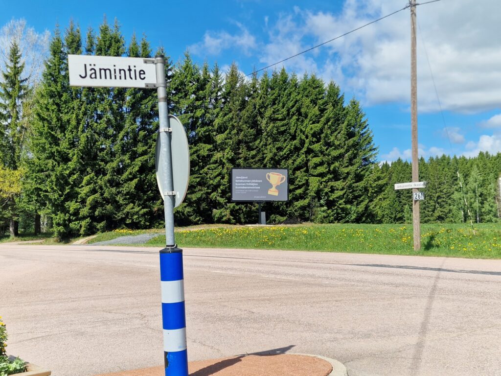 Digitaalinen infonäyttö ja siinä esitettävä mainos kuusiaidan edessä aurinkoisena kevätpäivänä.
