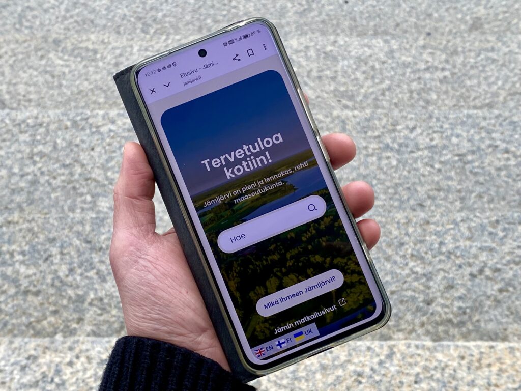 Kädessä puhelin, jossa näkyy jamijarvi.fi-sivuston etusivu ja kieliversioiden symbolit.