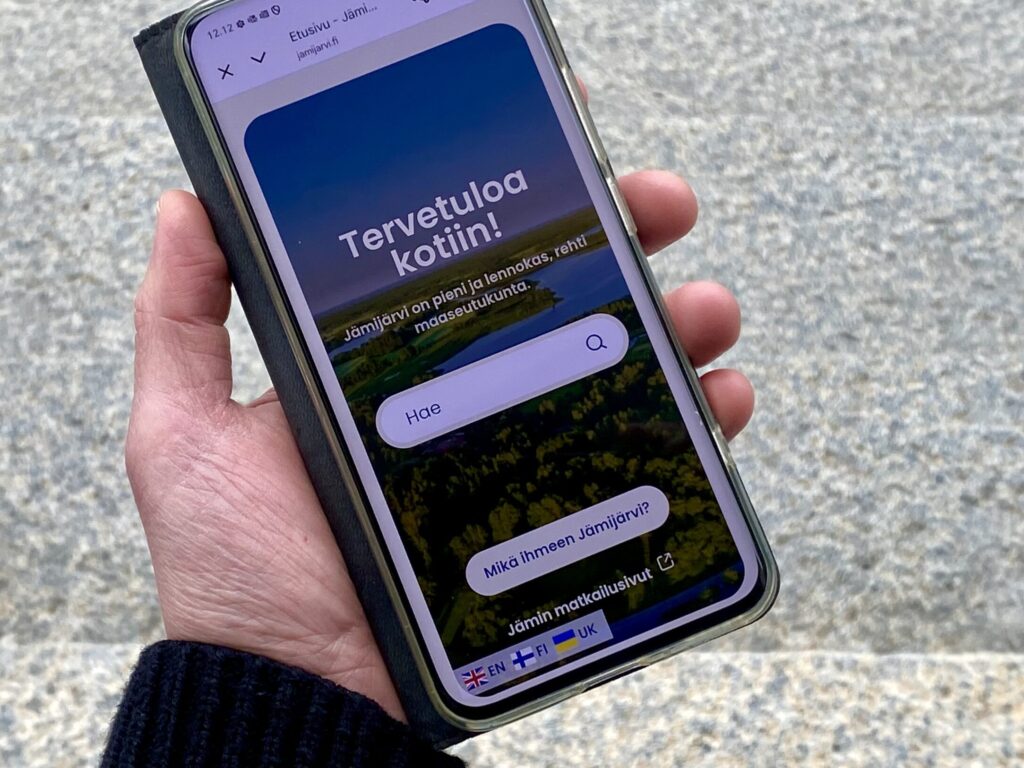 Kädessä puhelin, jossa näkyy jamijarvi.fi-sivuston etusivu ja kieliversioiden symbolit.
